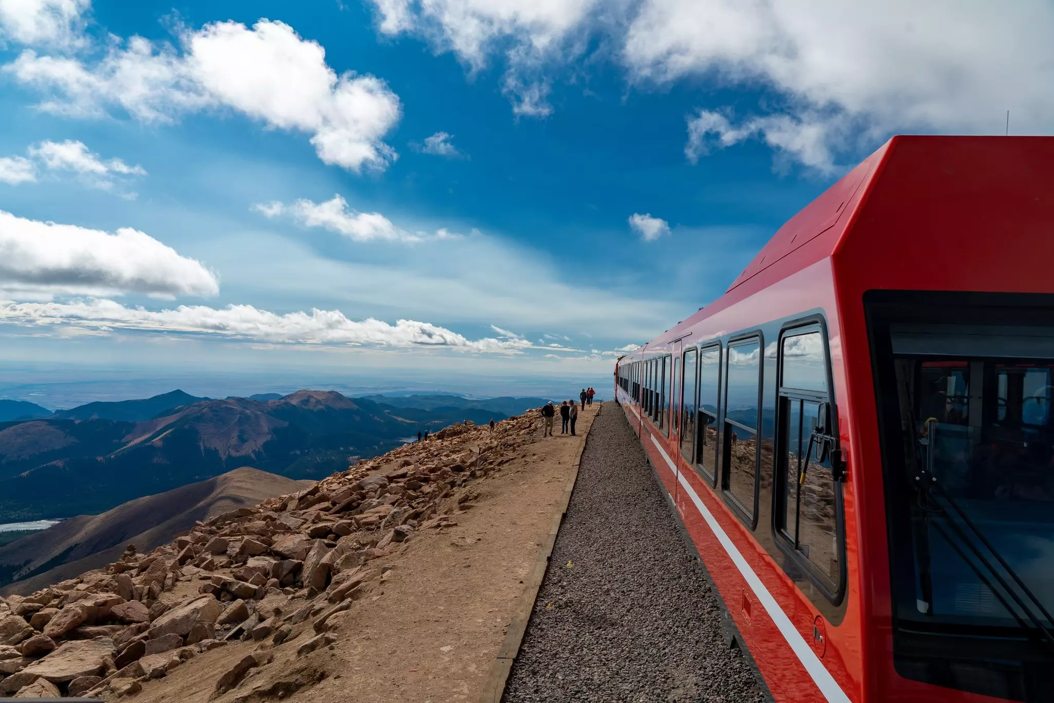 Red-painted train cars are stationed at the top of mountain, with passengers waiting to board along the edge of the track. Dramatic mountain views are visible below the train.