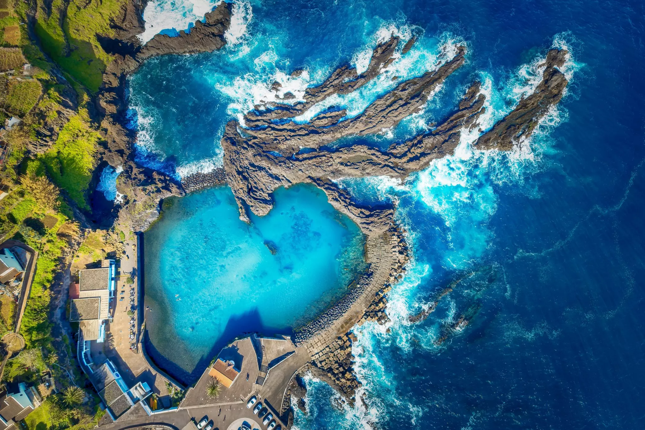 An aerial view of a bright blue natural pool carved from rock next to the rocky shore of the sea.
