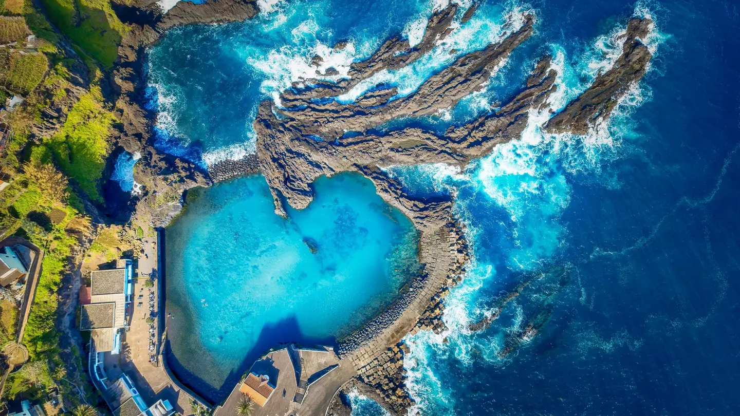 An aerial view of a bright blue natural pool carved from rock next to the rocky shore of the sea.