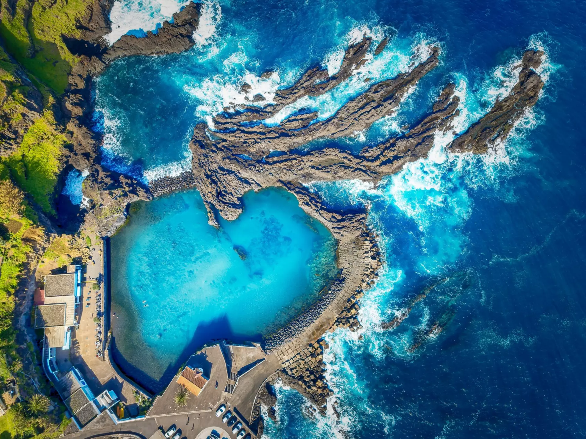 An aerial view of a bright blue natural pool carved from rock next to the rocky shore of the sea.