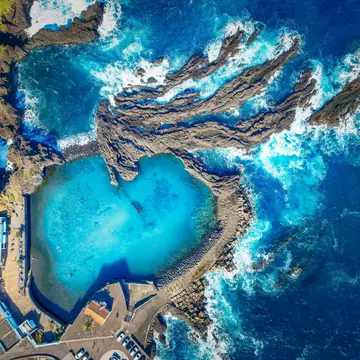 An aerial view of a bright blue natural pool carved from rock next to the rocky shore of the sea.
