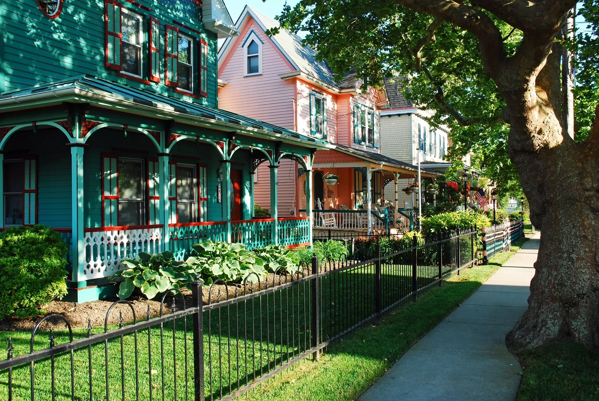 Colorful Victorian houses with wraparound porches line a tree-shaded sidewalk on a sunny street.
