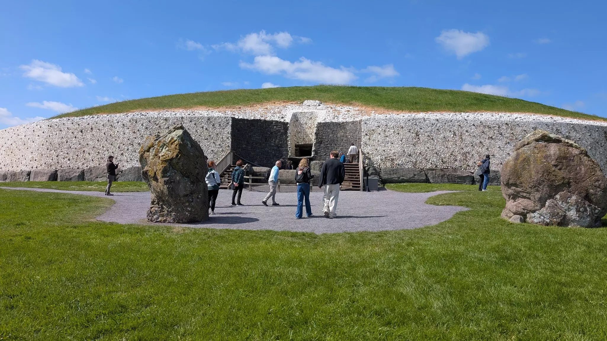 People walk around the outside of a large circular ancient tomb with huge stone boulders in the nearby grass.