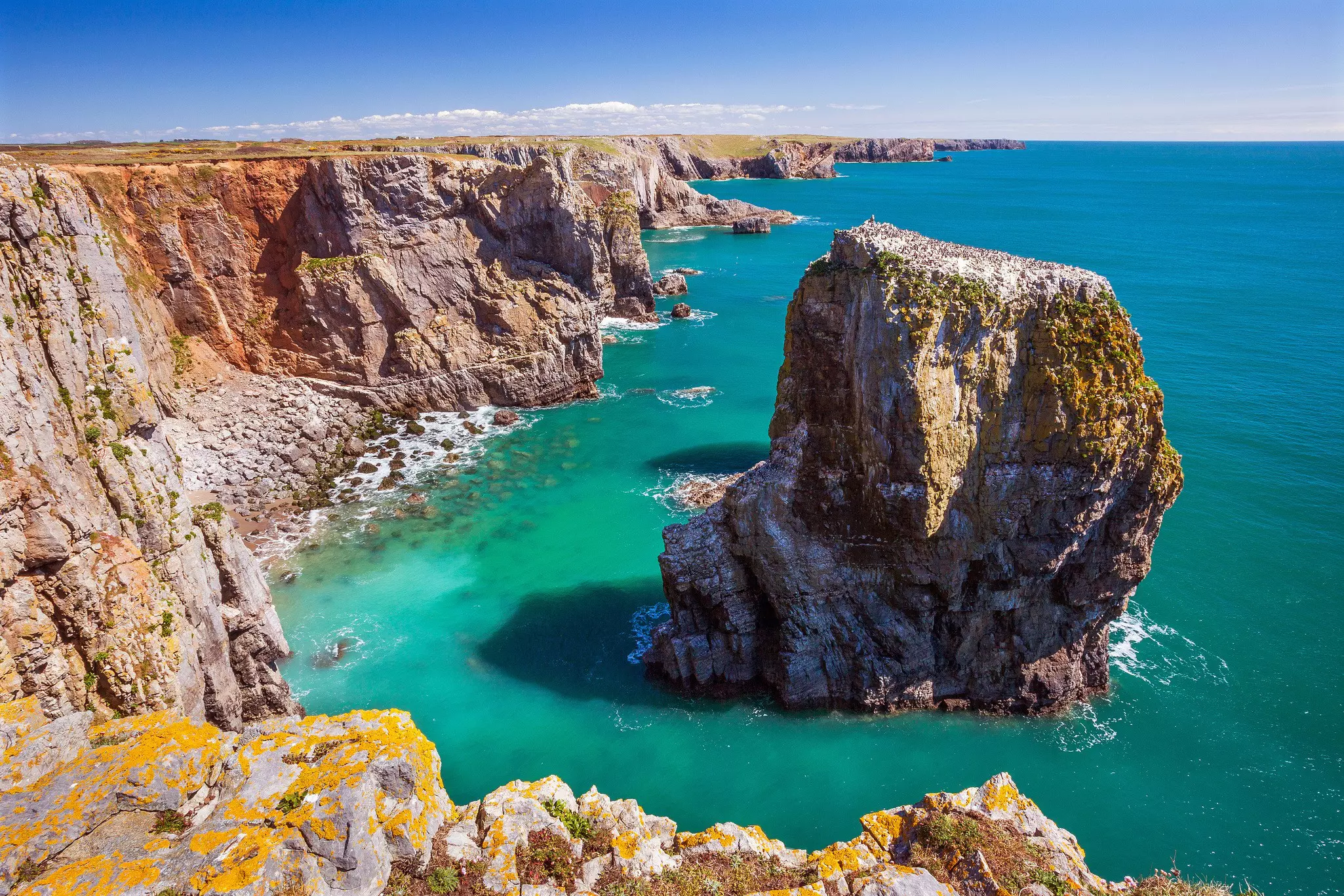 A rocky stack in the bright-blue sea is seen from the top of cliffs along a dramatic shoreline.