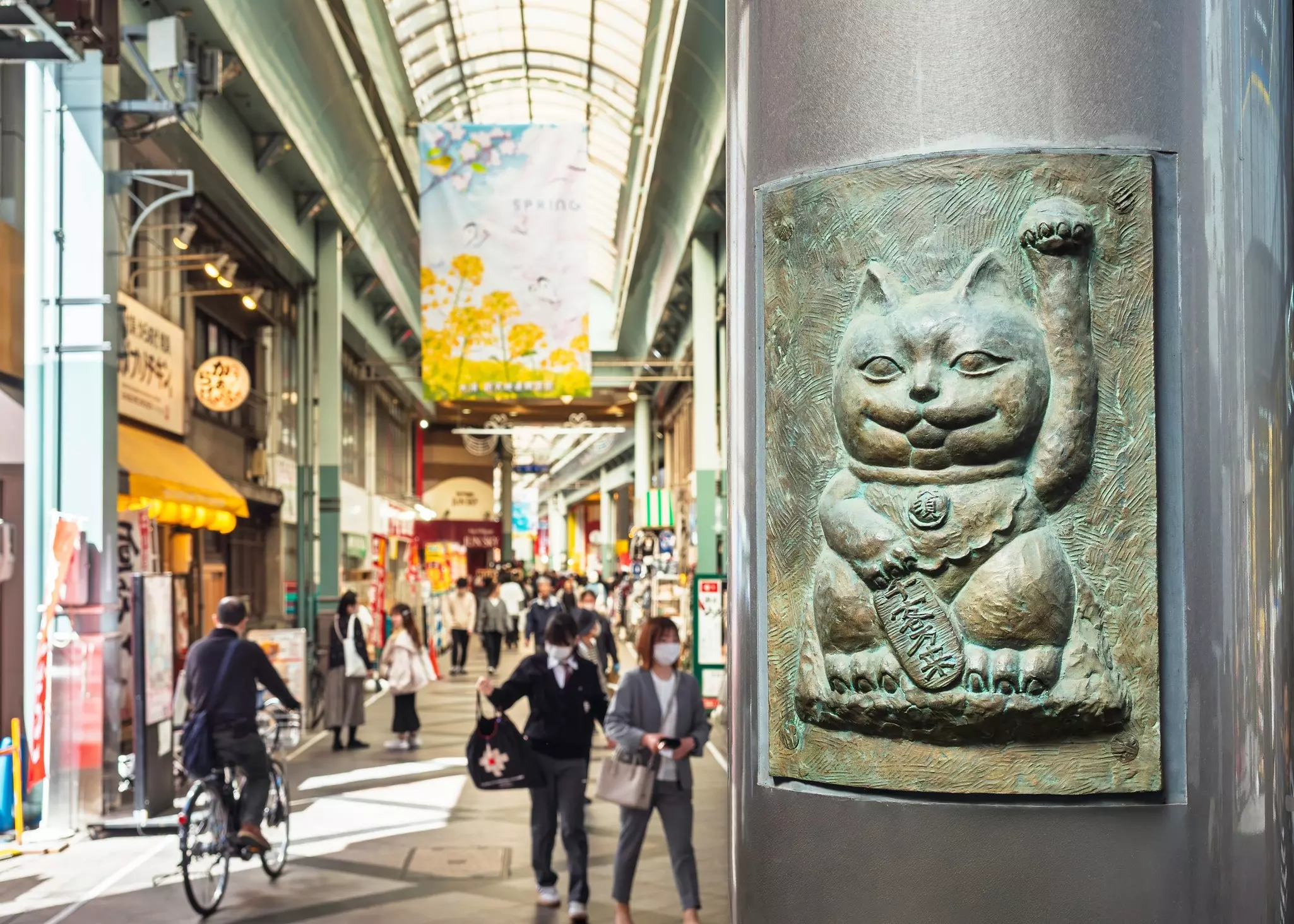 Pedestrians and a man riding a bike pass through a covered street lined with shops. A large relief of a waving cat decorates a nearby column.