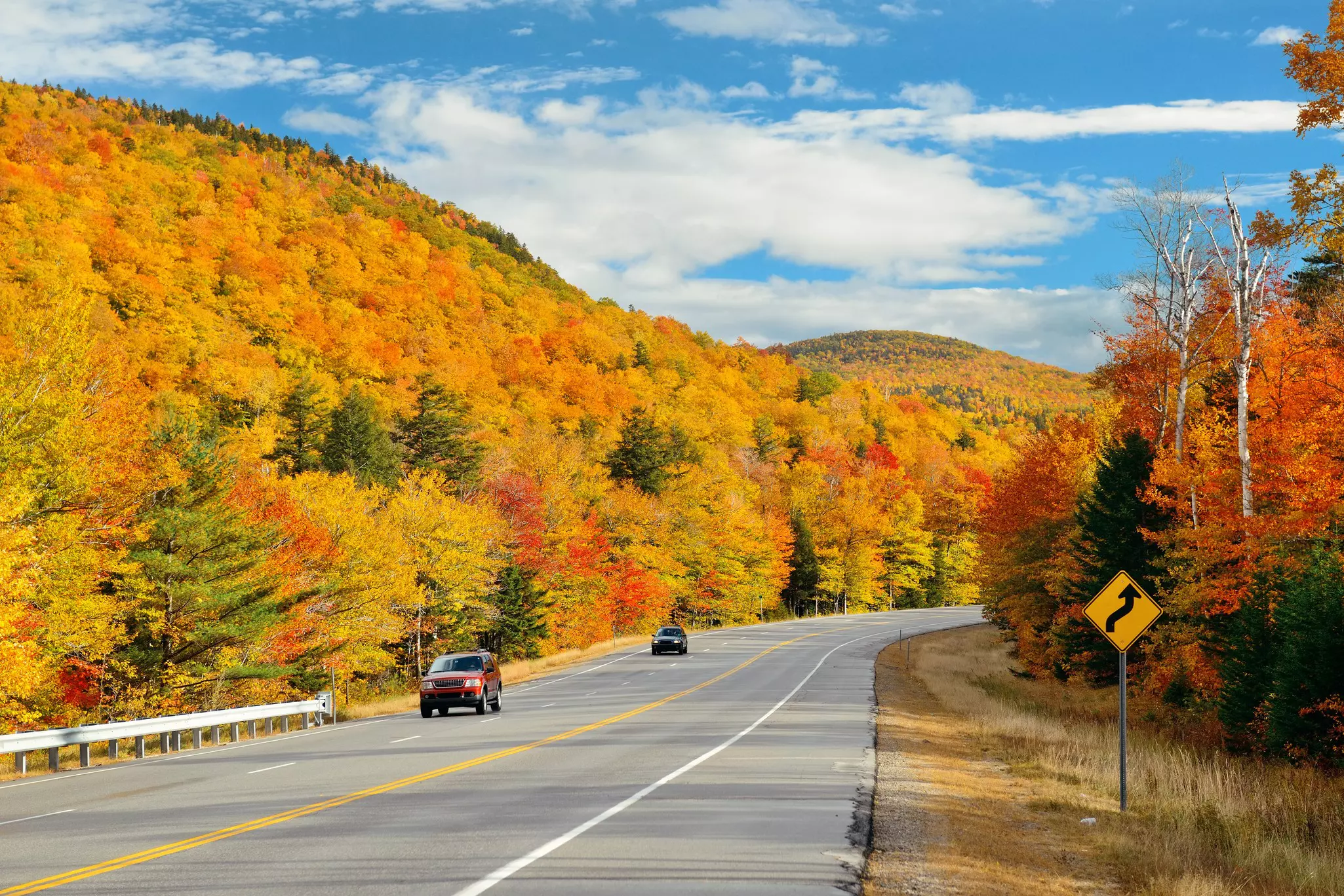 Two cars drive on a three-lane road, with trees in autumn colors on either side and a blue sky with white clouds above.