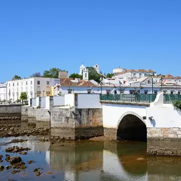 A white bridge over shallow water with white buildings in the background