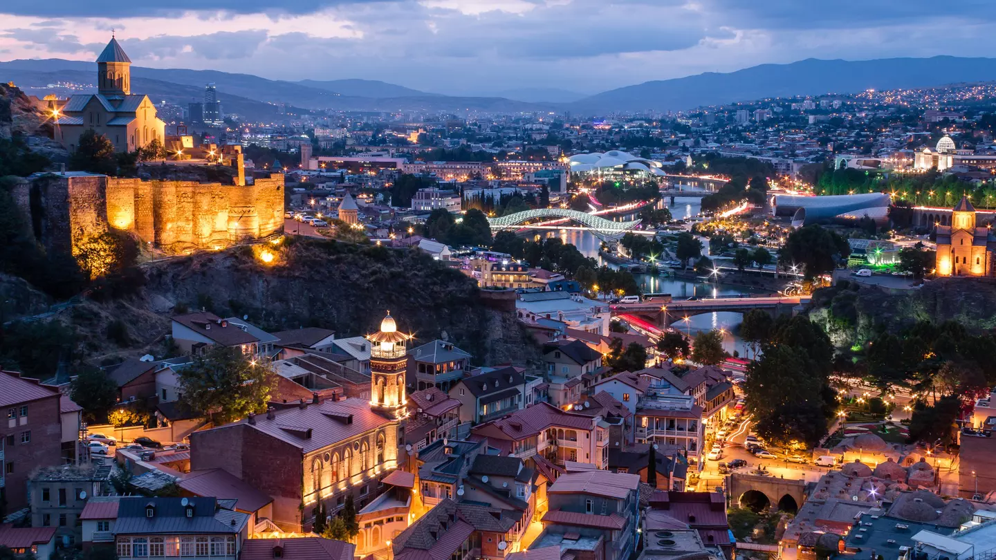City view at dusk. A hilltop church with fortress-like walls is lit up to the left. There is a river with two illuminated bridges.