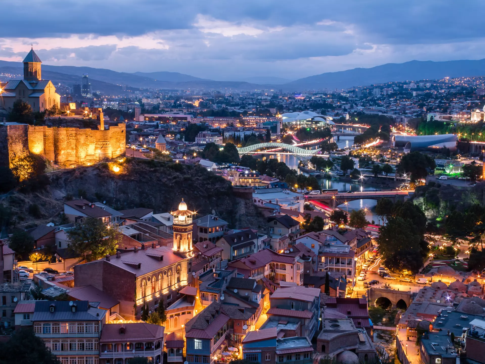 City view at dusk. A hilltop church with fortress-like walls is lit up to the left. There is a river with two illuminated bridges.