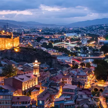 City view at dusk. A hilltop church with fortress-like walls is lit up to the left. There is a river with two illuminated bridges.