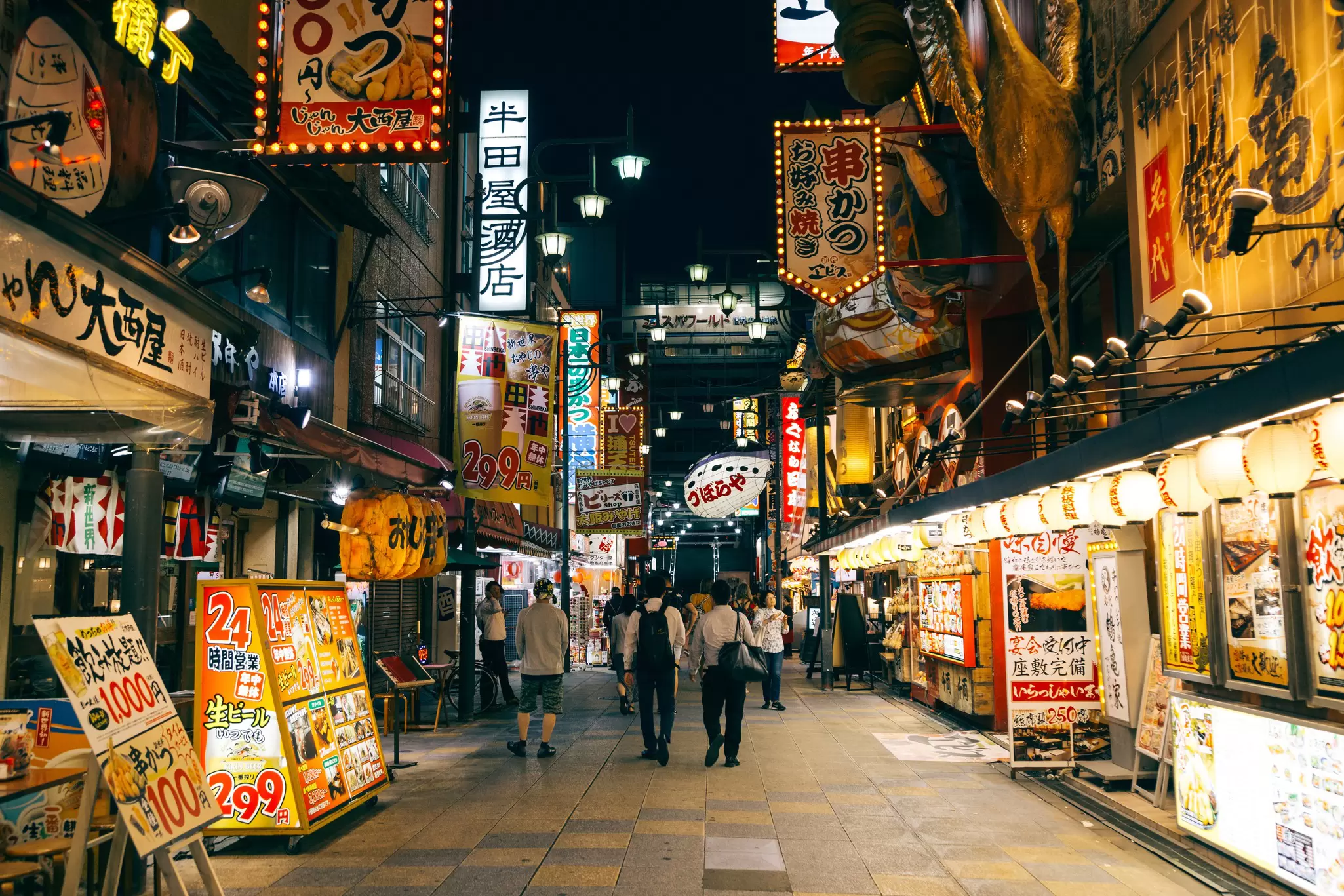 Brightly lit restaurants at night lining a street in the Shinsekai district of Osaka.
