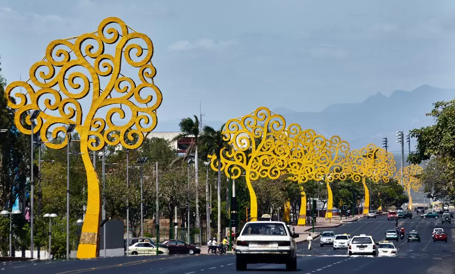 Giant yellow metal Trees of Life sculptures lining a busy avenue in Managua, Nicaragua, with taxis and hills beyond