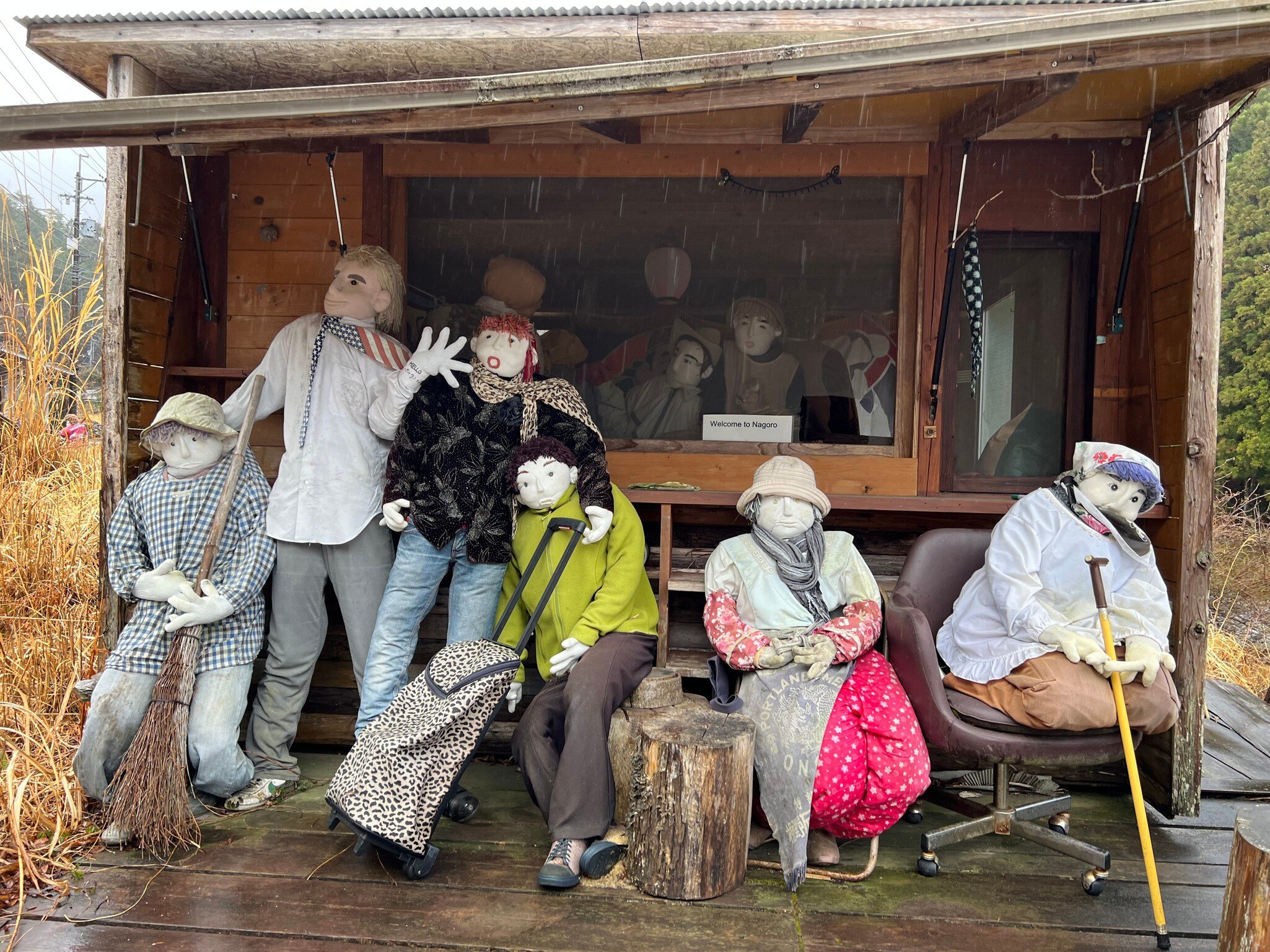 Six life-size cloth dolls sit and stand on a rainy wooden veranda; a small sign reads "Welcome to Nagoro".
