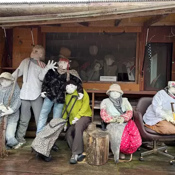 Six life-size cloth dolls sit and stand on a rainy wooden veranda; a small sign reads "Welcome to Nagoro".