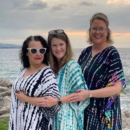Three friends in flowy colorful tops pose for a photo by the sea at sunset
