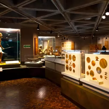 A museum gallery displays gold objects in cases, as well as life-size dioramas of Indigenous people.