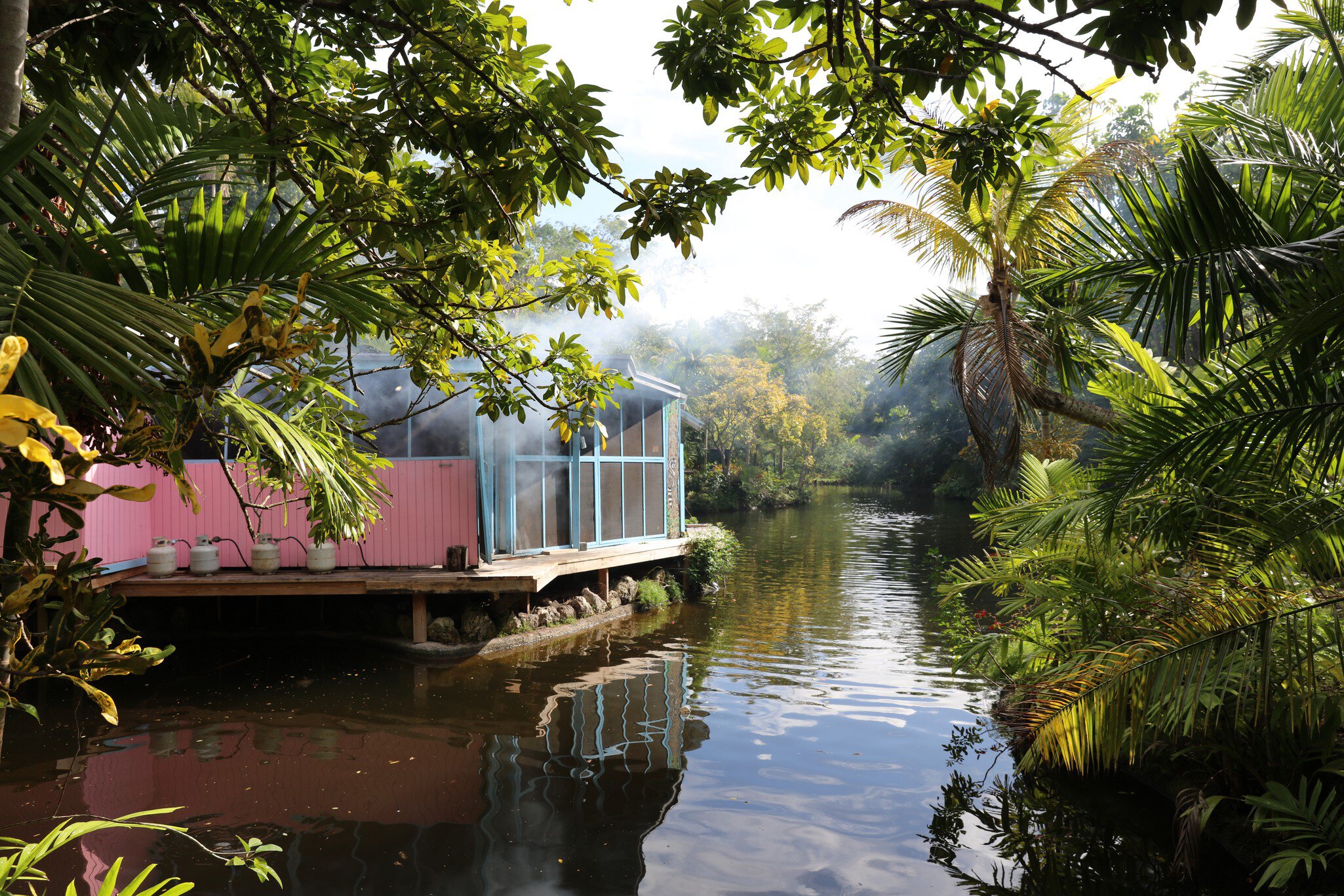 Pink and blue waterside cabin with propane tanks reflected in a calm tropical river fringed by palms and foliage