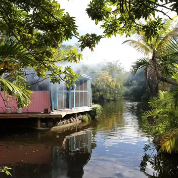 Pink and blue waterside cabin with propane tanks reflected in a calm tropical river fringed by palms and foliage