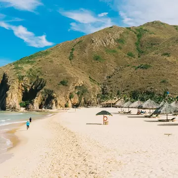 White-sand beach with thatched umbrellas and loungers, two people walking by the water below a green hill, Ky Co, Quy Nhon.