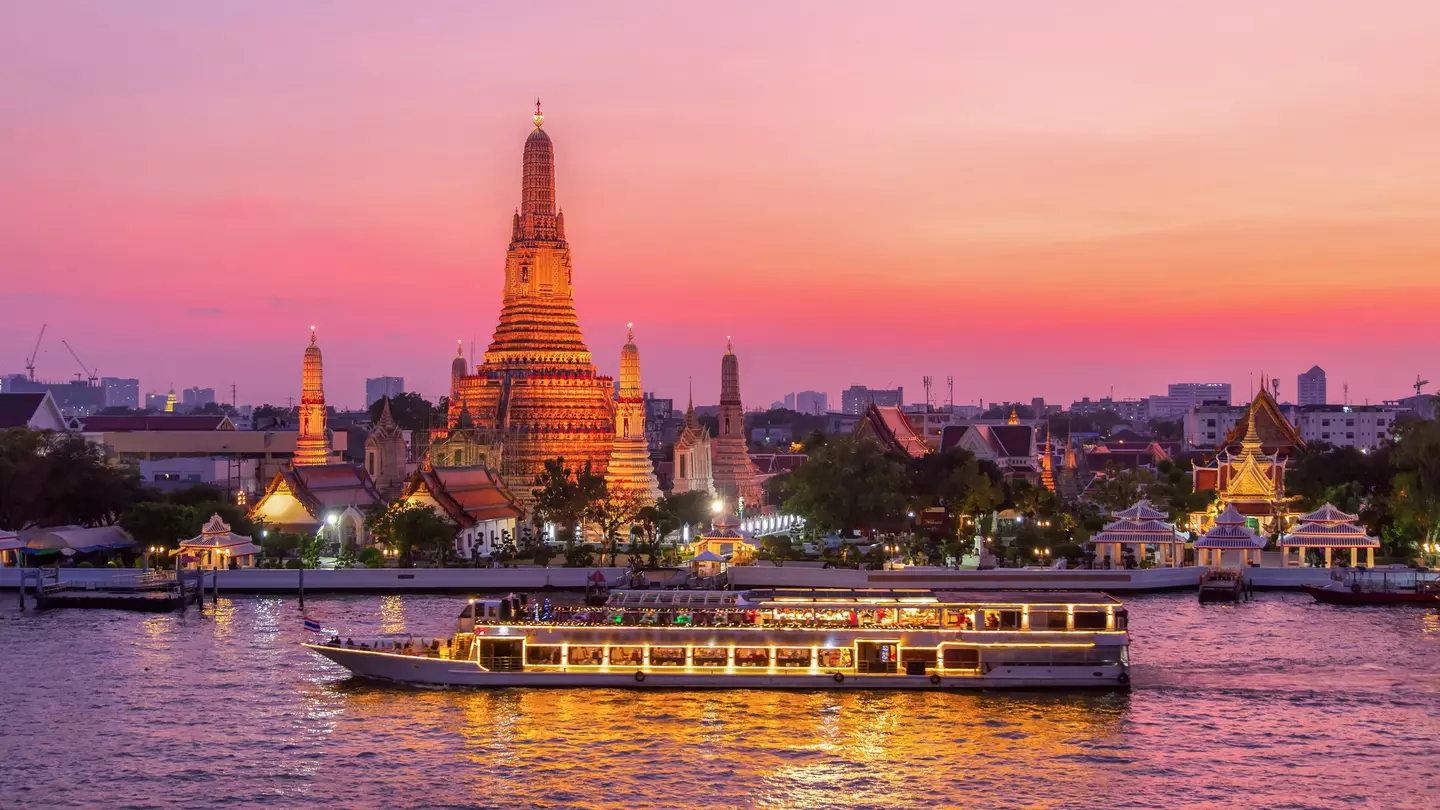 A boat with lights on its windows passes a tall stupa by a river. The scene is bathed with pink and orange sunset light.