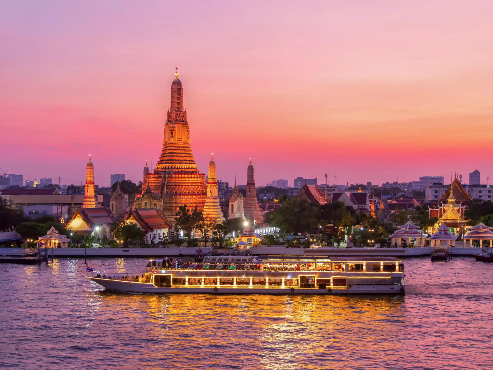 A boat with lights on its windows passes a tall stupa by a river. The scene is bathed with pink and orange sunset light.