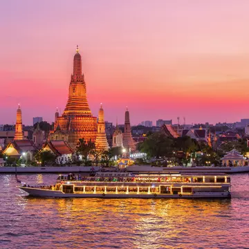 A boat with lights on its windows passes a tall stupa by a river. The scene is bathed with pink and orange sunset light.