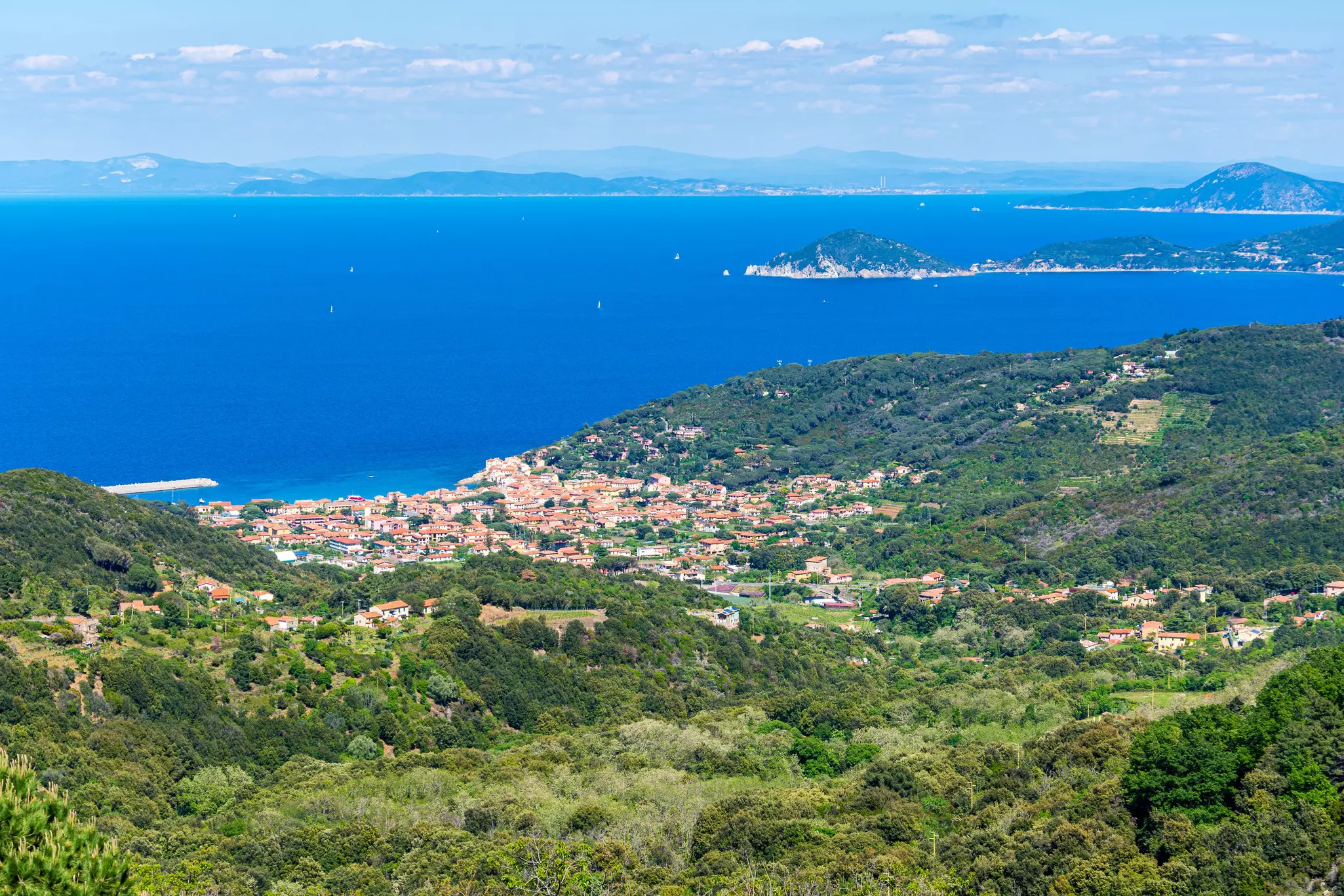 Aerial shot of green hills, red-topped houses in a seaside village, and the ocean and hills in the distance.