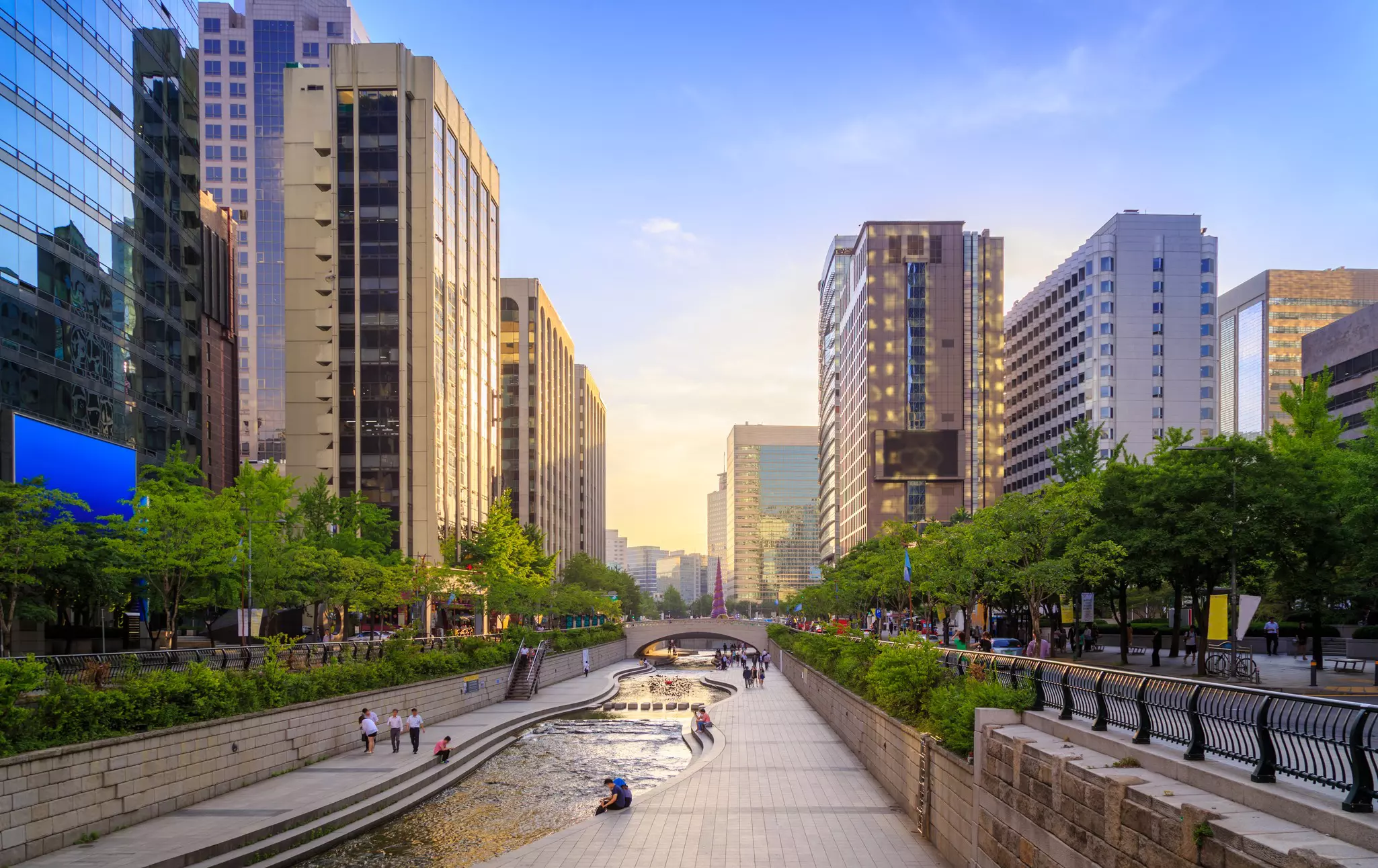 A stream running through the center of a high-rise city, with people sat on the banks, chatting and relaxing.