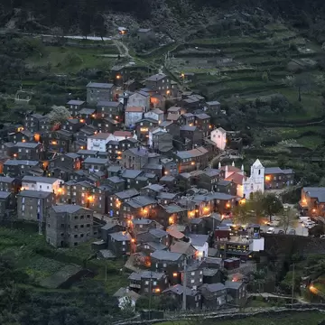 Dark schist houses of Piódão village, Portugal, with lit windows and a white church, on a terraced green hillside at dusk.