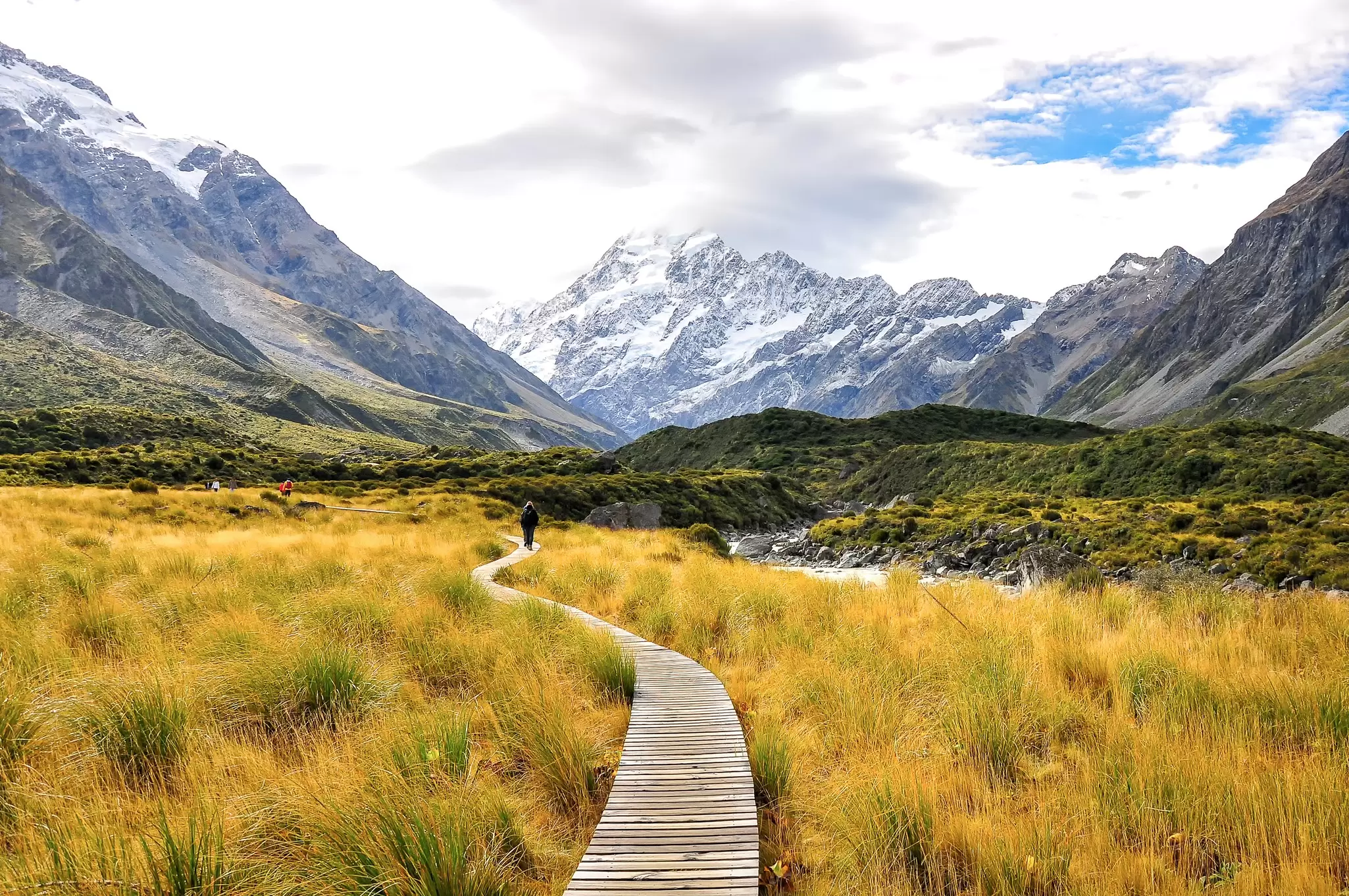 A hiker on a wooden walkway across green and yellow grass with mountains in the background.