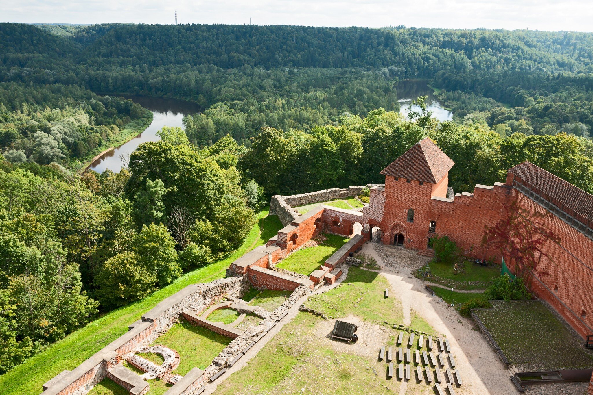 Aerial view of a red-brick castle and stone ruins on a wooded hill above a river.