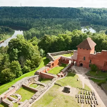 Aerial view of a red-brick castle and stone ruins on a wooded hill above a river.