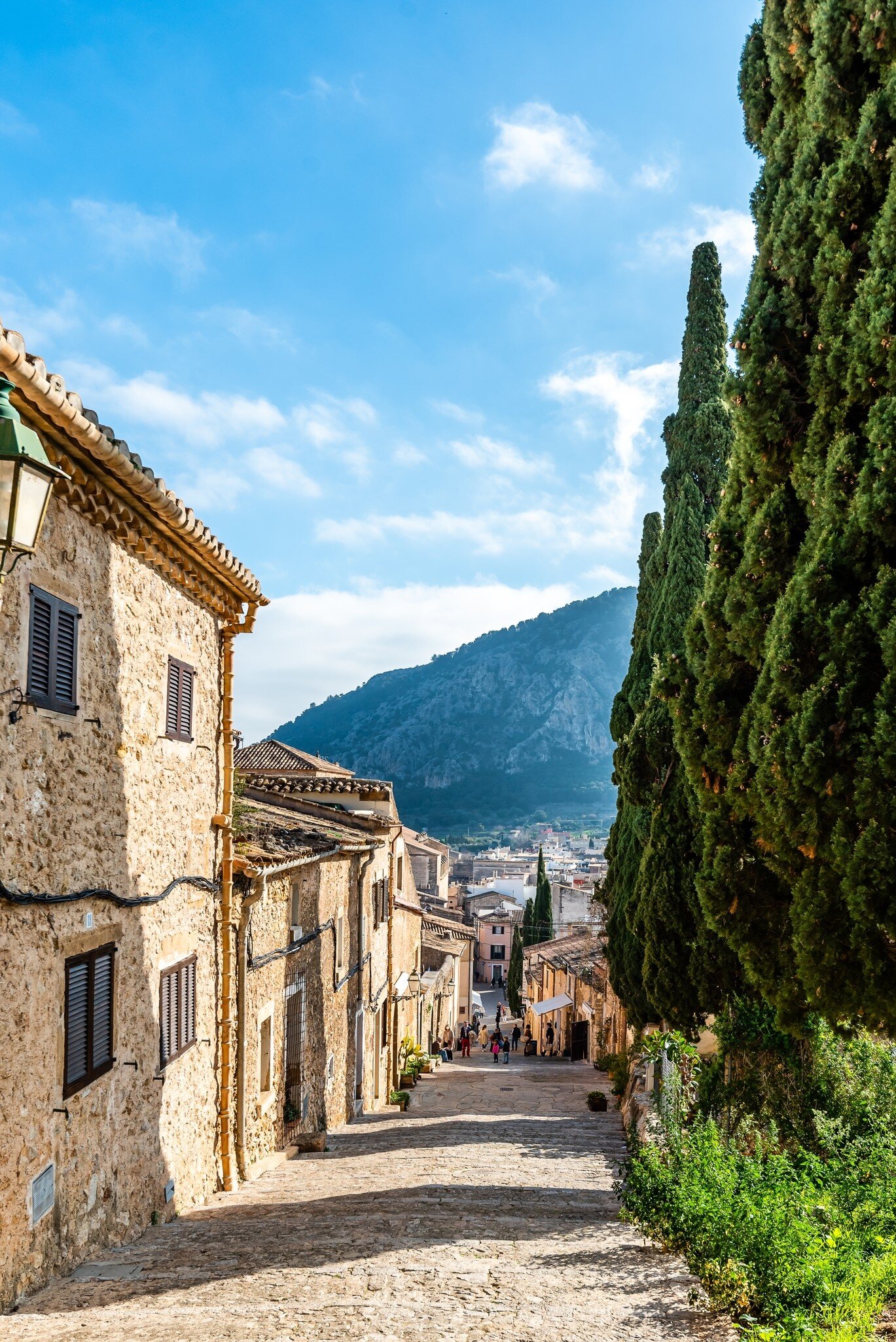 A sloping cobbled street of old stone houses leading down toward a mountain in a Mediterranean village.