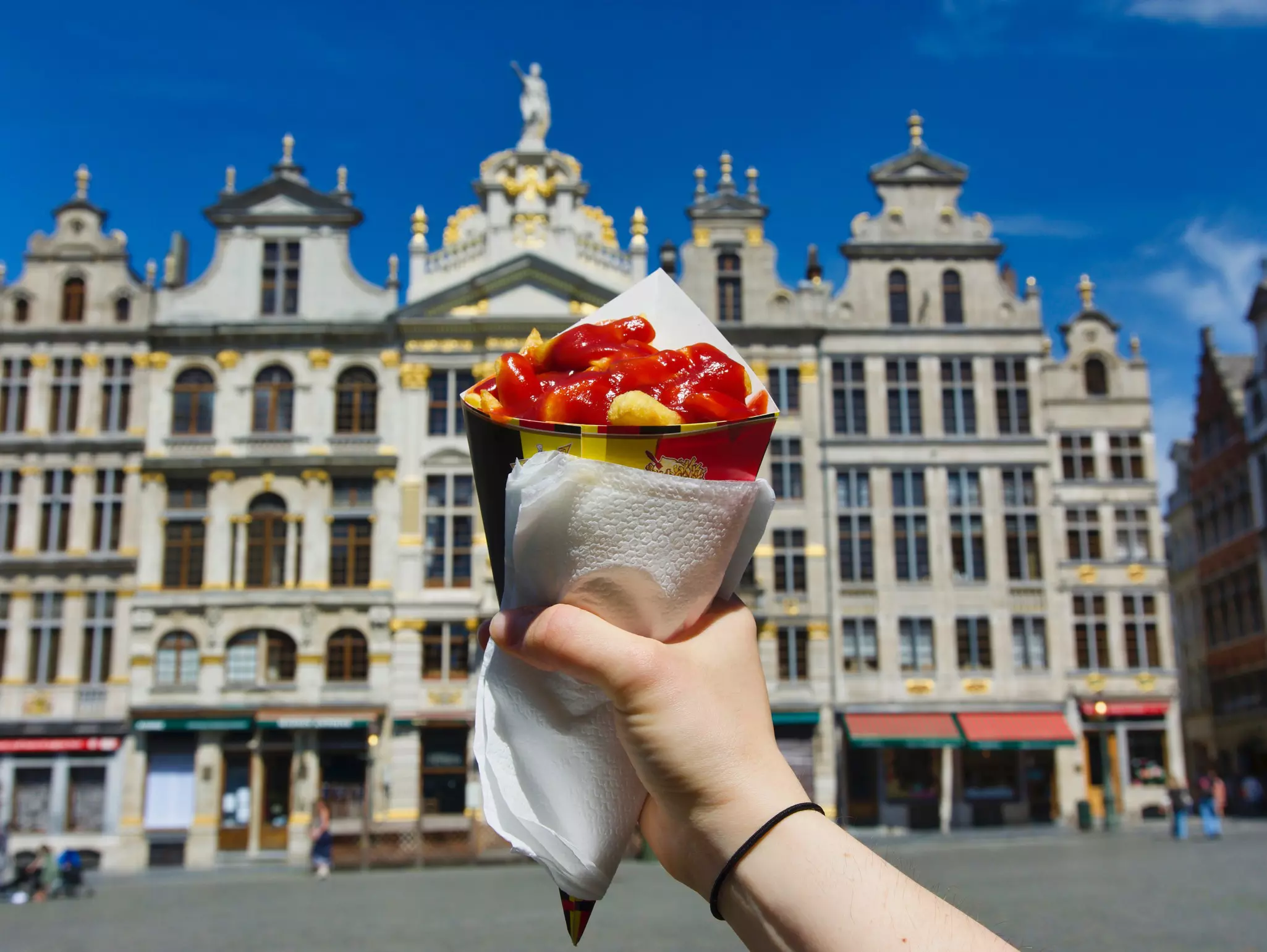 A hand holds up a cone of Belgian fries in front of an ornate building in Brussels