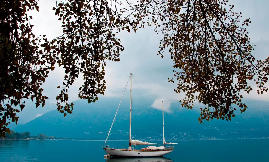 Two-masted sailboat moored on Lake Maggiore near Locarno, Switzerland, framed by overhanging branches and misty mountains