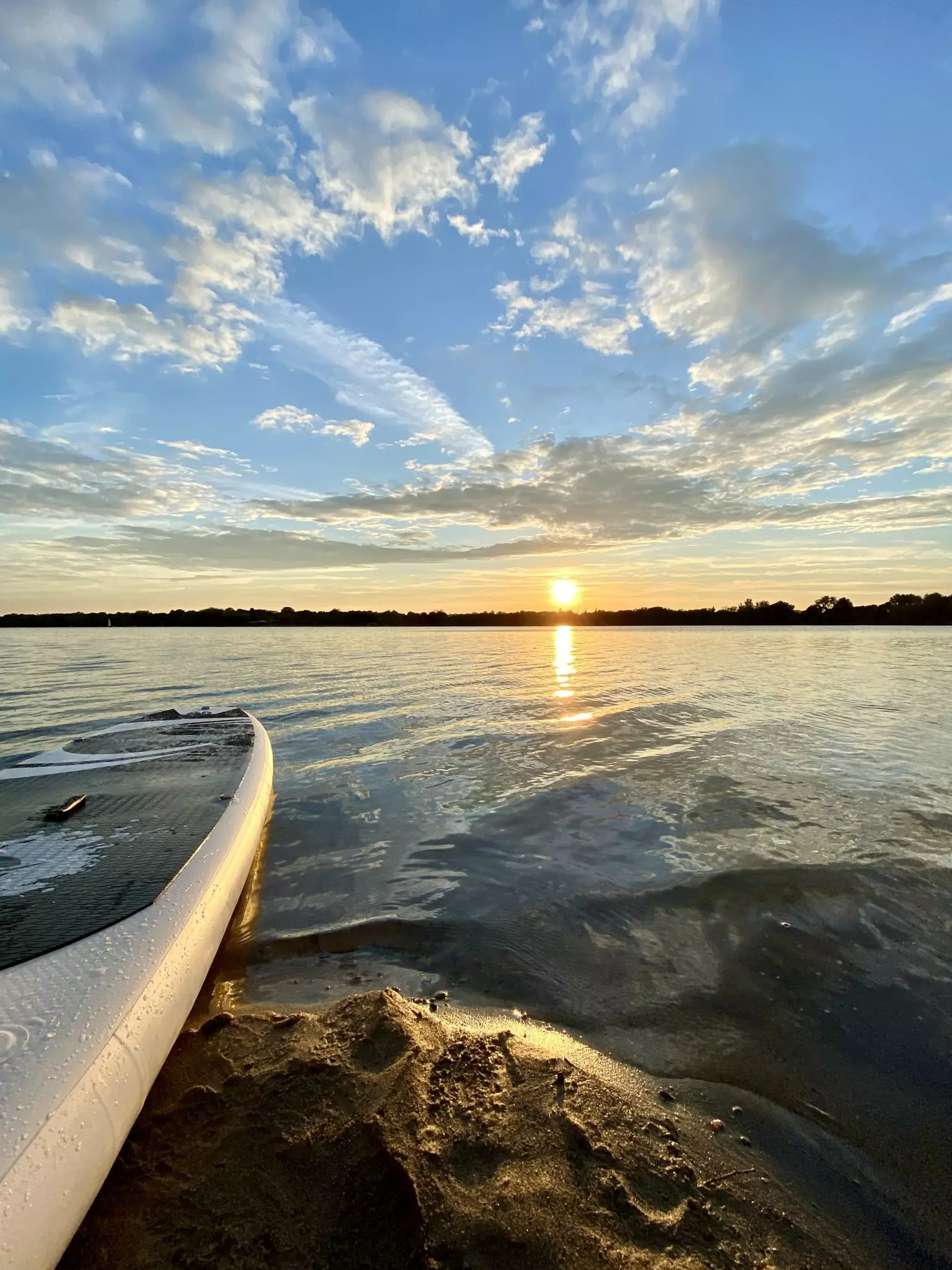 A shot looking down a paddleboard out onto a lake as the sun sends orange streaks across a blue cloudy sky