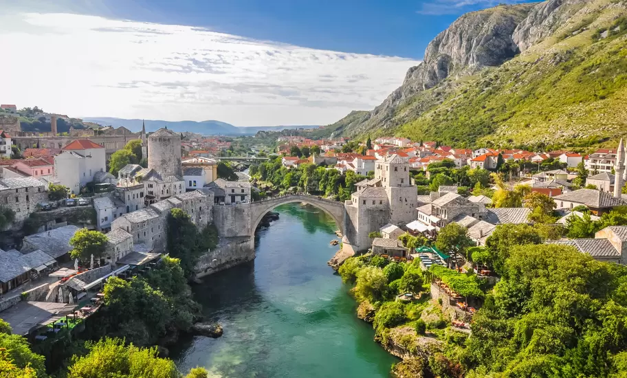 Mostar's Stari Most stone bridge spanning the green Neretva River, with old town houses, minarets and a mountain behind
