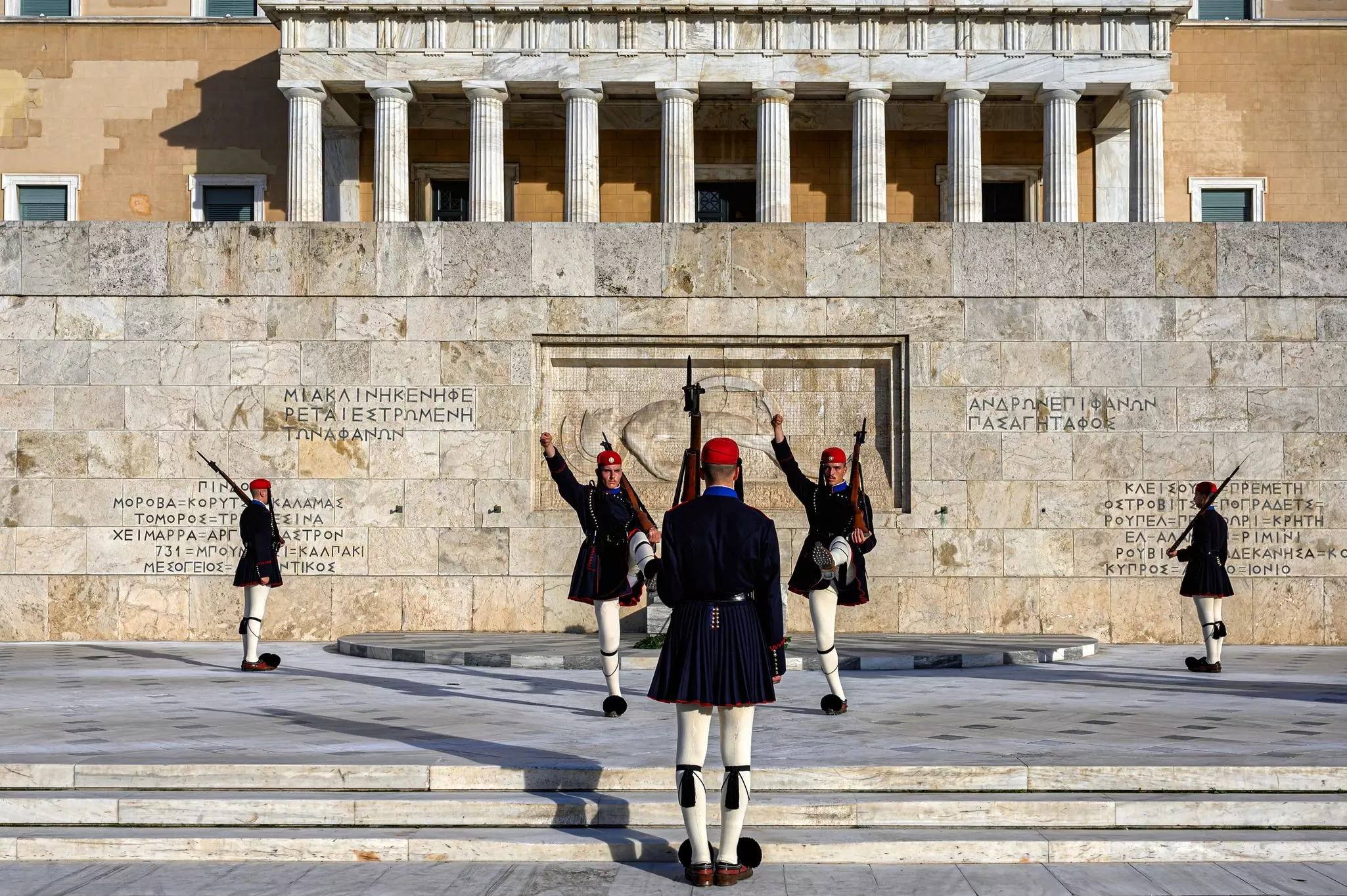 Five guards outside a building with Greek writing on its walls. One is at the center with his back to the camera, facing two guards whose right arms and left legs are raised. The other two guards are on either side of the frame, in profile view.