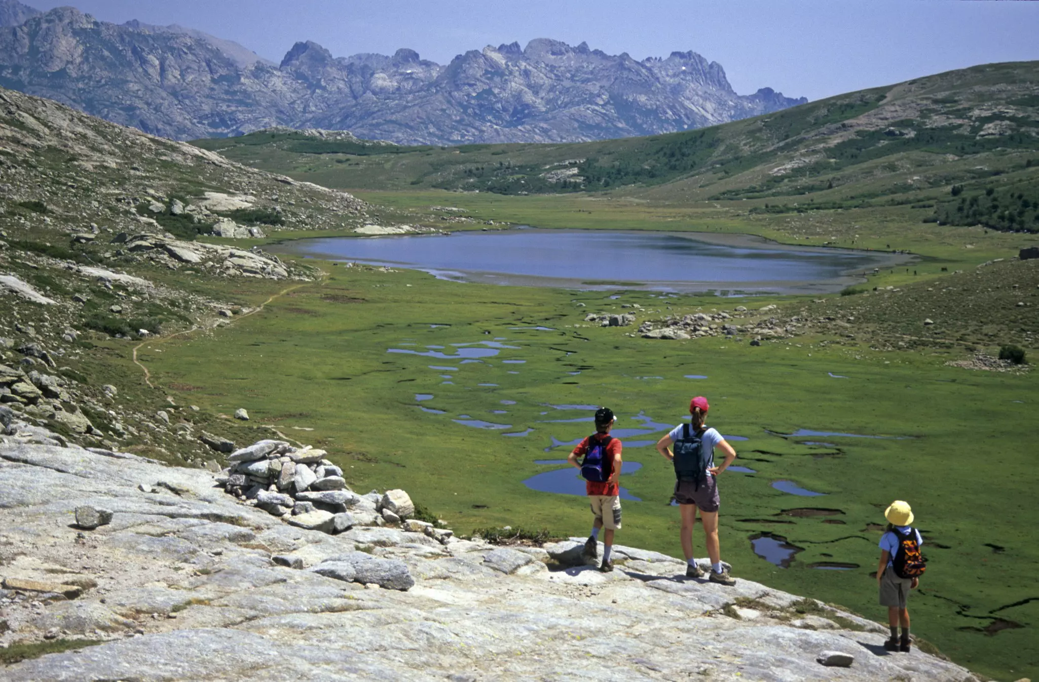 A few hikers pause on a rock overlooking lush grassland dotted with small ponds heading towards mountains.