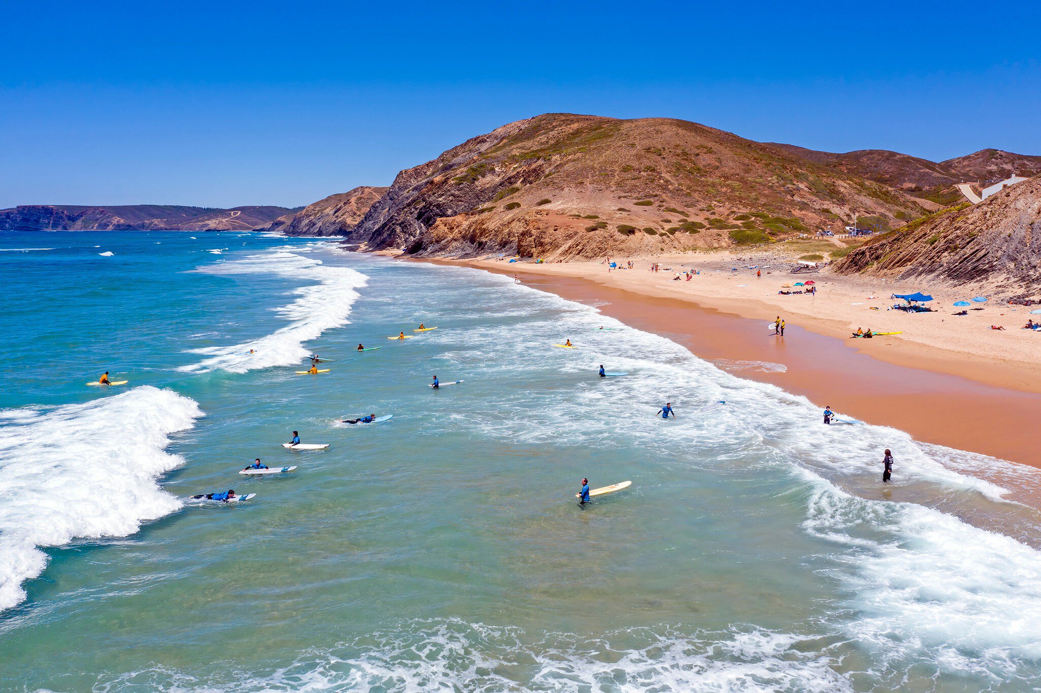 Surfers waiting in the waters off a rocky red coastline.