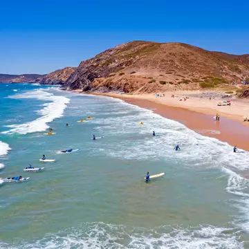Surfers waiting in the waters off a rocky red coastline.