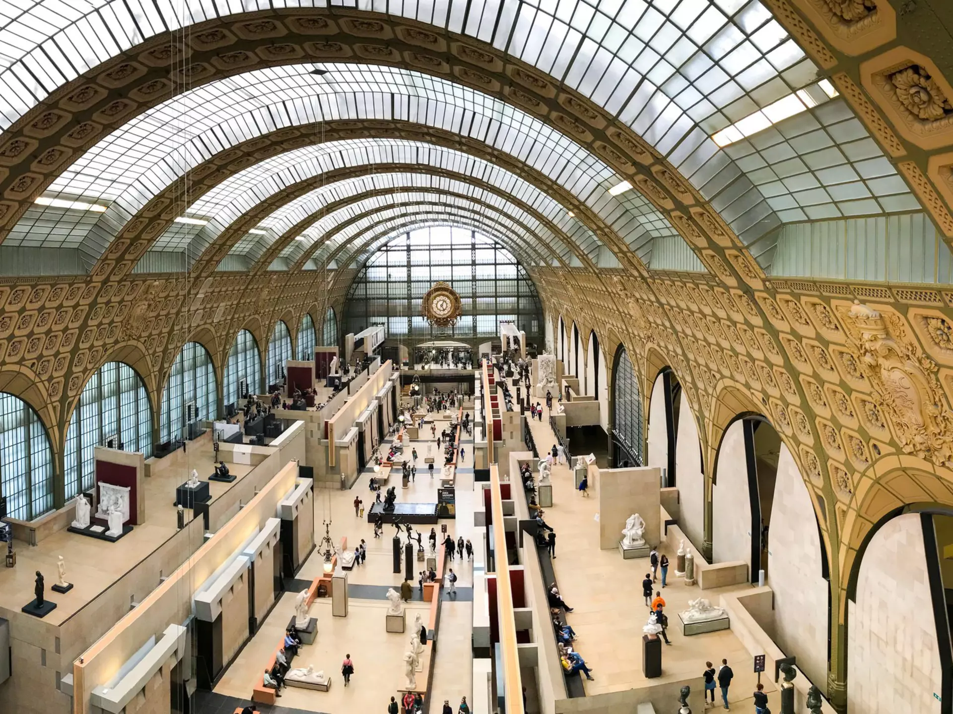 People move around sculptures on display in a vast exhibition space in a former train station with galleries to the side.