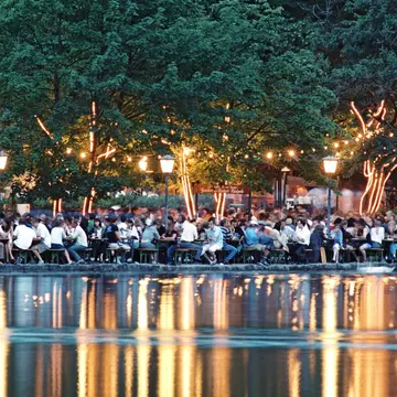 A pool table sits under a crystal chandelier in front of a deep purple wall decorated with a colorful illustration of tropical plants. A white midcentury modern chair sits in the corner and white French doors sit at the far end of the pool table