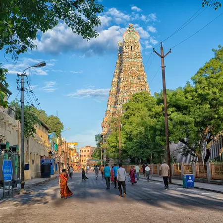 People walk down a street that's overlooked by a tall temple tower with ornate sculptures on its facade