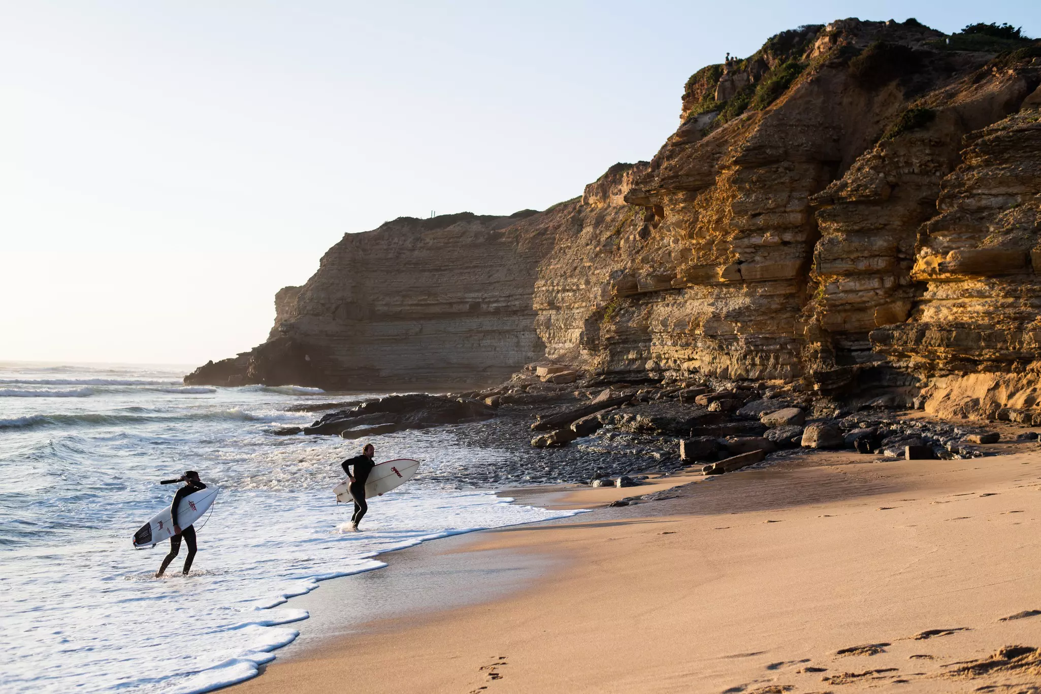 Surfers emerge from the waves onto the sandy beach at Praia da Ribeira d'Ilhas, Portugal. 
