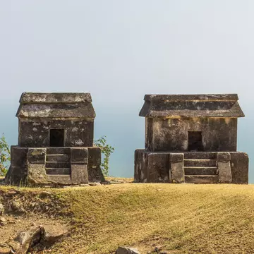 Ancient mausoleum-style tombs on a grassy hillside above the sea on a mostly sunny day.