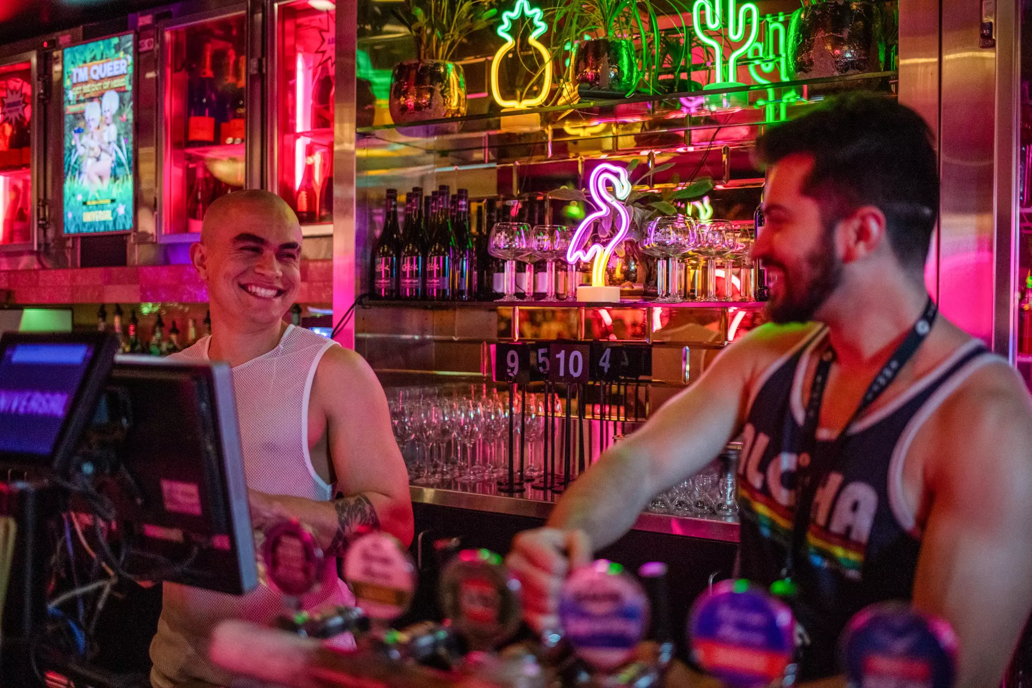 Two bartenders stand behind a bar. Neon signs and bottles line the wall behind them.
