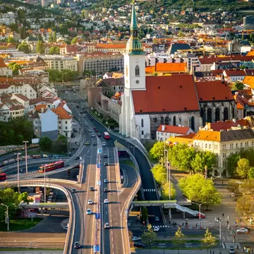 Gothic cathedral with a green spire and gold crown above red rooftops, a bridge and river in the foreground.