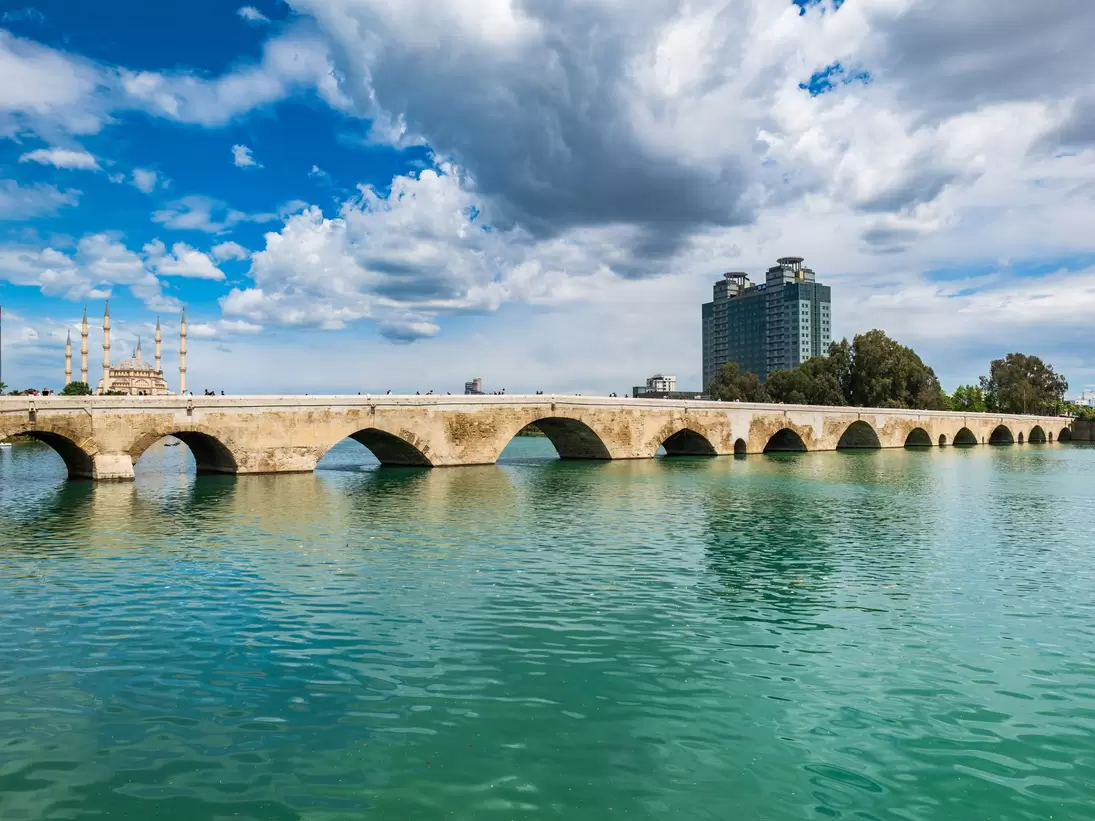 A long arched stone bridge over a green river in Adana, Turkey, with mosque minarets and a tower block under a cloudy sky.