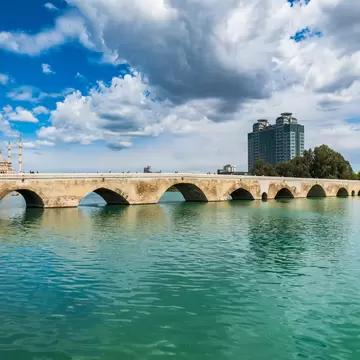A long arched stone bridge over a green river in Adana, Turkey, with mosque minarets and a tower block under a cloudy sky.